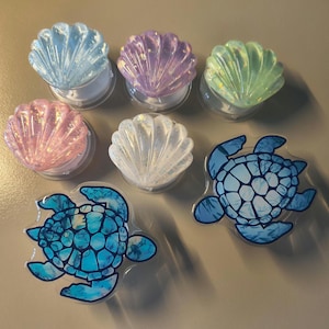 May include: A selection of mobile phone grips with seashell and sea turtle designs. The seashell grips are in blue, purple, green, pink, and white. The sea turtle grips are blue with a mosaic pattern.