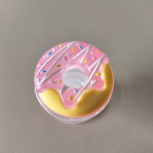May include: A pink donut-shaped container with white icing and colourful sprinkles. The bottom half is yellow, and the container is clear. The design resembles a frosted donut.
