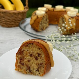 May include: A slice of banana bread on a white plate, topped with a caramel-colored glaze and a banana slice. Other banana bread pieces are on a glass plate, garnished with banana slices and chocolate chips. A basket of bananas is in the background.