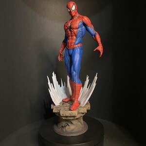 Pode incluir: Uma figura detalhada do Homem-Aranha numa pose din&acirc;mica, mostrando seu ic&oacute;nico fato vermelho e azul. A figura est&aacute; sobre uma base rochosa com efeitos de teia de aranha brancos estilizados. A est&aacute;tua tem cerca de 30 cm de altura.