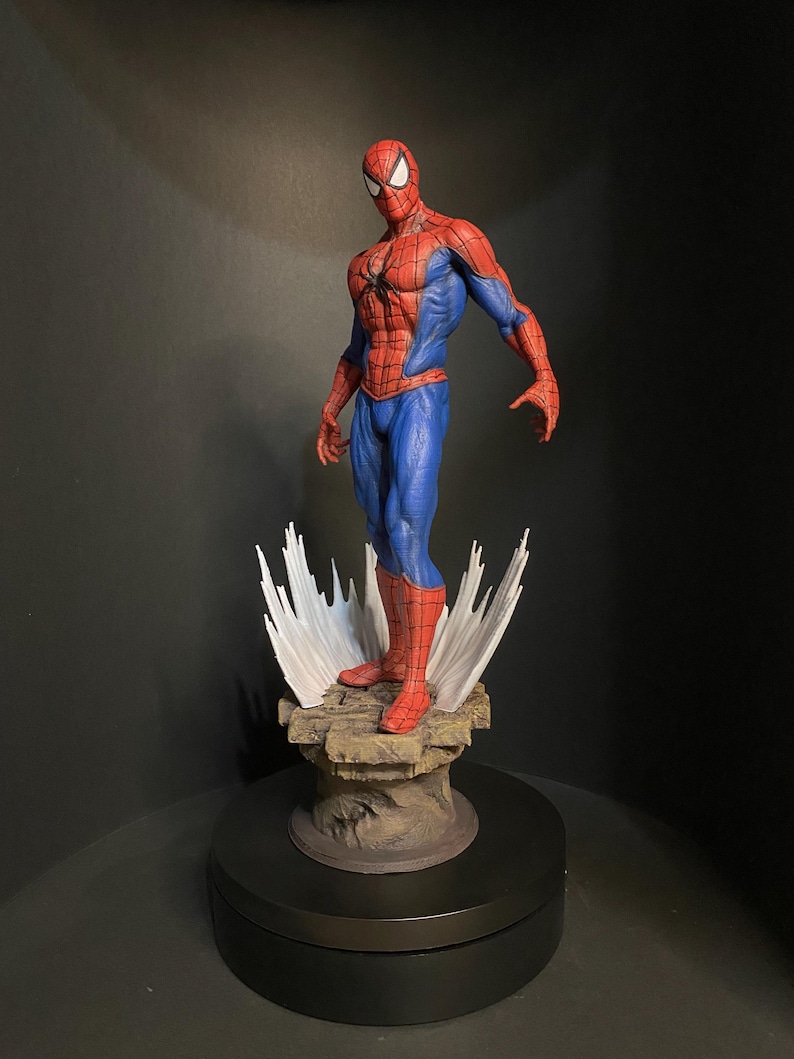 Pode incluir: Uma figura detalhada do Homem-Aranha numa pose din&acirc;mica, mostrando seu ic&oacute;nico fato vermelho e azul. A figura est&aacute; sobre uma base rochosa com efeitos de teia de aranha brancos estilizados. A est&aacute;tua tem cerca de 30 cm de altura.