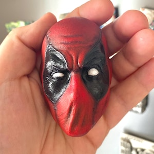 May include: A red and black Deadpool mask figurine with white eyes. The mask is held in a hand, showcasing the detailed paintwork and design. The figurine is a collectible item, likely for fans of the Deadpool comic books and movies.