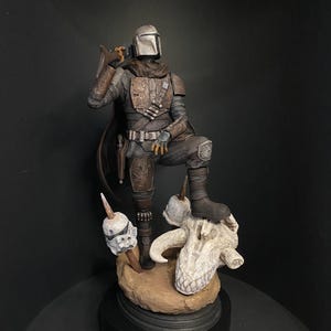 May include: A detailed figurine of The Mandalorian in a standing pose, wearing his signature armor and helmet. The figure is posed with one foot on a skull and a blaster in hand. The base is a dark brown color.