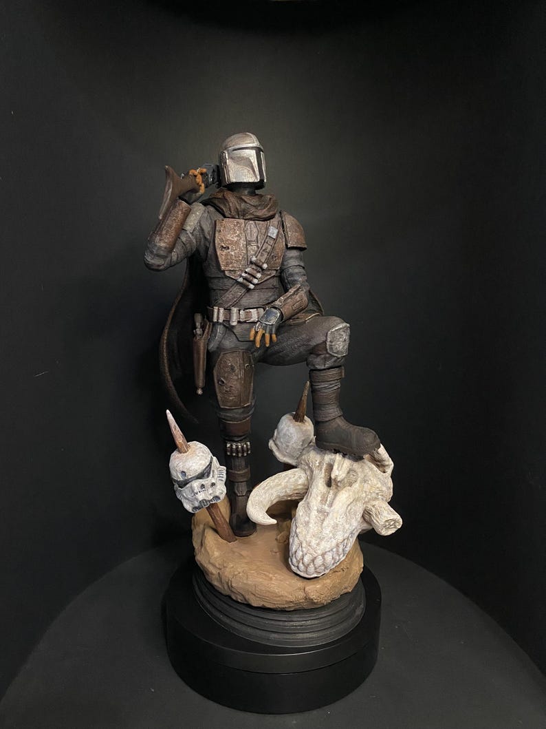 May include: A detailed figurine of The Mandalorian in a standing pose, wearing his signature armor and helmet. The figure is posed with one foot on a skull and a blaster in hand. The base is a dark brown color.