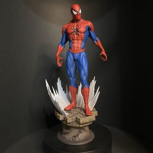 Pode incluir: Figura detalhada do Homem-Aranha em pose cl&aacute;ssica, vestindo um traje vermelho e azul com um emblema de aranha preta no peito. A figura est&aacute; sobre uma base rochosa com efeitos de teia de aranha brancos estilizados.