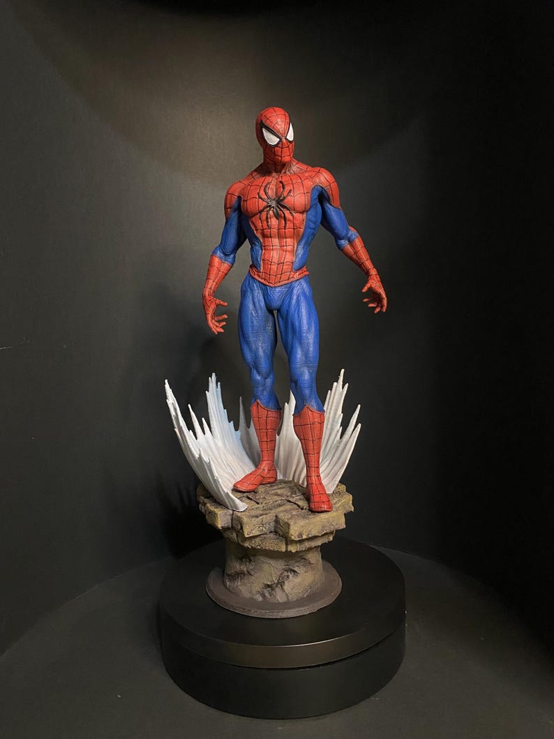 Pode incluir: Figura detalhada do Homem-Aranha em pose cl&aacute;ssica, vestindo um traje vermelho e azul com um emblema de aranha preta no peito. A figura est&aacute; sobre uma base rochosa com efeitos de teia de aranha brancos estilizados.