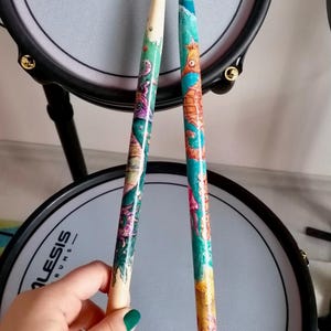Puede incluir: Dos baquetas de batería de madera con diseños coloridos de criaturas marinas. Las baquetas están pintadas con tonos de turquesa, naranja y morado, y se muestran frente a una almohadilla de batería gris. Una persona con esmalte de uñas verde sostiene las baquetas.