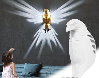 May include: A golden eagle-shaped wall lamp with outstretched wings, casting a radiant light pattern. The lamp is mounted on a dark gray wall. A white, geometric bird sculpture is also visible.
