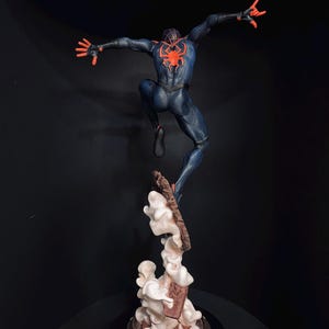 Puede incluir: Figura detallada de un superh&eacute;roe en una pose din&aacute;mica, saltando por el aire. La figura est&aacute; vestida con un traje azul oscuro con un emblema de ara&ntilde;a roja en la espalda y detalles rojos. La base presenta un dise&ntilde;o estilizado de nubes y escombros.