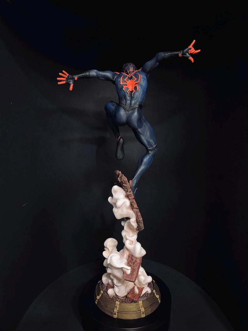 Puede incluir: Figura detallada de un superh&eacute;roe en una pose din&aacute;mica, saltando por el aire. La figura est&aacute; vestida con un traje azul oscuro con un emblema de ara&ntilde;a roja en la espalda y detalles rojos. La base presenta un dise&ntilde;o estilizado de nubes y escombros.