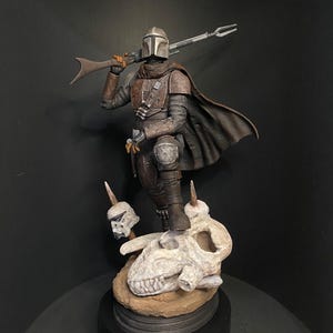 May include: A detailed figurine of The Mandalorian, a character from the Star Wars franchise, standing on a skull base. The figure is clad in brown and gray armor, holding a rifle. The base features a skull and two smaller skull figures.