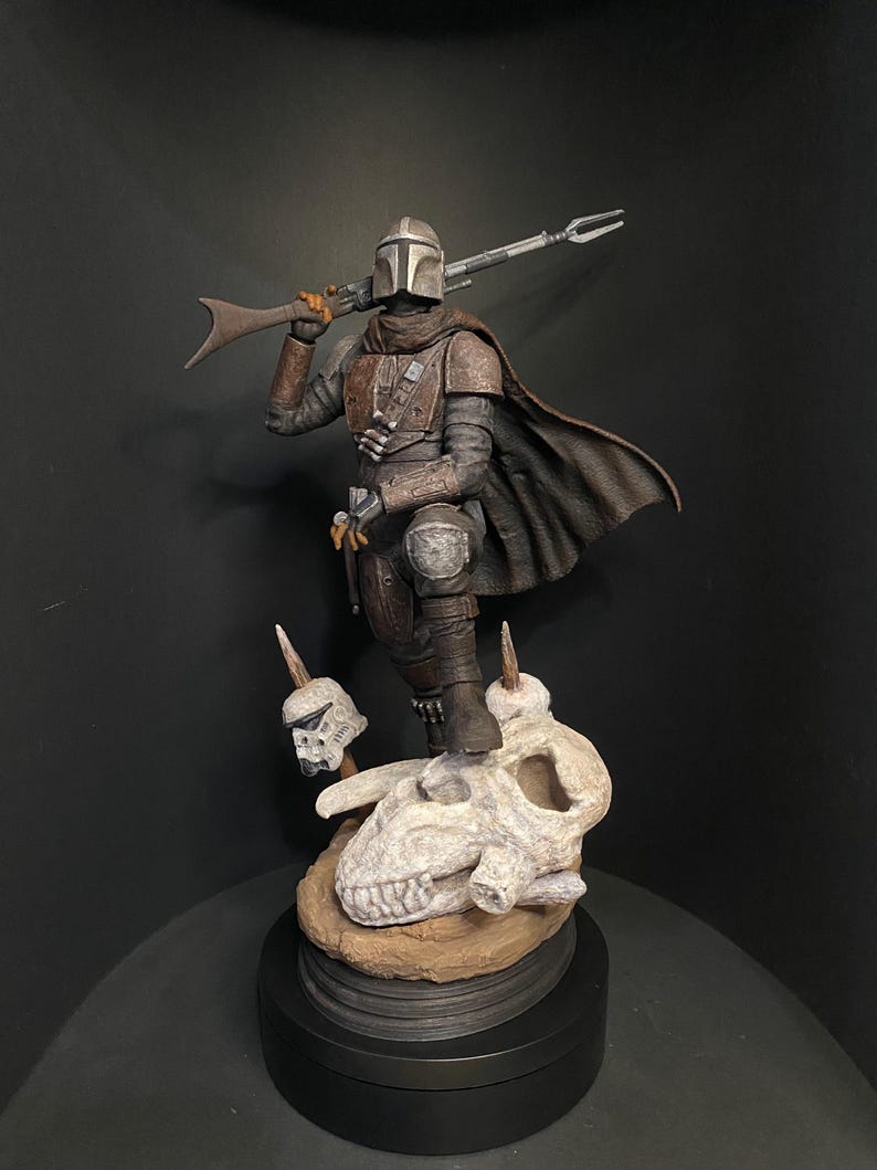 May include: A detailed figurine of The Mandalorian, a character from the Star Wars franchise, standing on a skull base. The figure is clad in brown and gray armor, holding a rifle. The base features a skull and two smaller skull figures.