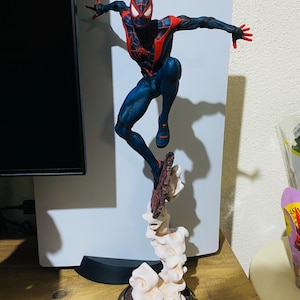 Puede incluir: Figura detallada de un superh&eacute;roe en una pose din&aacute;mica, con un traje negro y rojo con detalles de telara&ntilde;a. La figura est&aacute; en el aire, sostenida por una base estilizada con efectos de humo, sobre una superficie de madera.