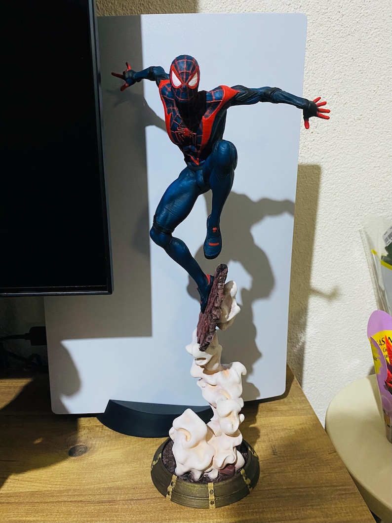 Puede incluir: Figura detallada de un superh&eacute;roe en una pose din&aacute;mica, con un traje negro y rojo con detalles de telara&ntilde;a. La figura est&aacute; en el aire, sostenida por una base estilizada con efectos de humo, sobre una superficie de madera.