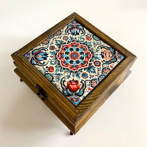 May include: A square wooden box with a decorative floral tile top. The tile features a detailed pattern in red, blue, and white. The box has a dark wood finish and small feet.