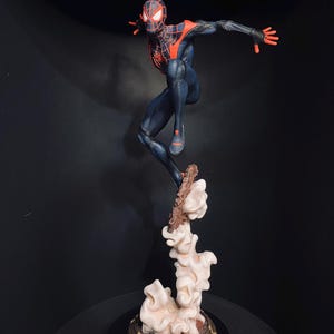 Puede incluir: Figura detallada de un superh&eacute;roe en una pose din&aacute;mica, vestido con un traje negro y rojo. La figura est&aacute; en el aire, sostenida por una base estilizada de nubes y escombros. La base es de dise&ntilde;o circular con detalles dorados. La m&aacute;scara del superh&eacute;roe tiene ojos blancos brillantes.
