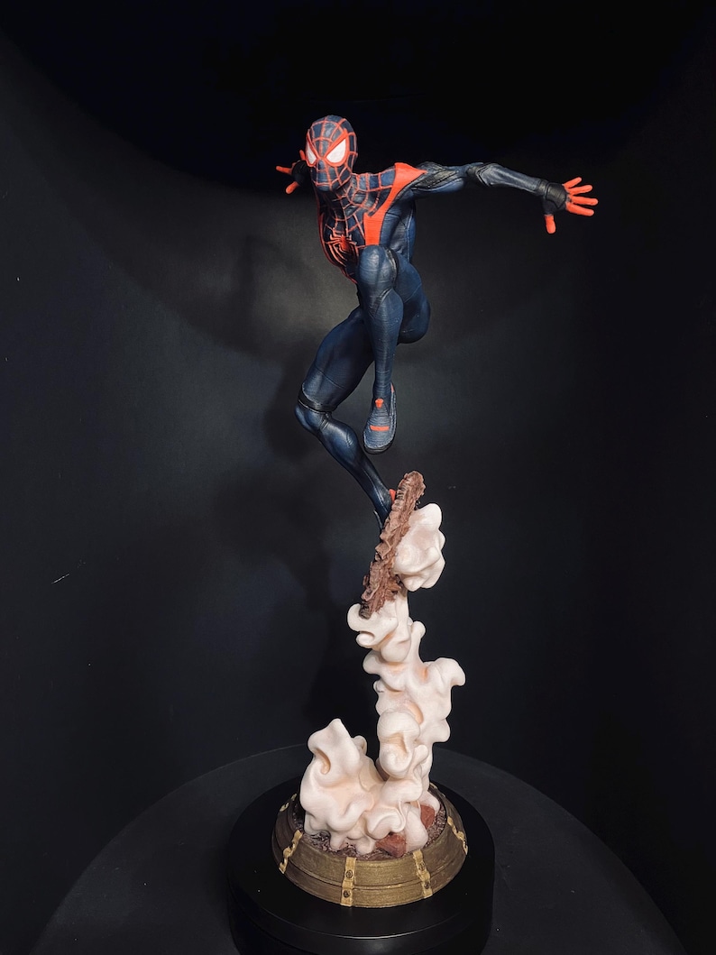 Puede incluir: Figura detallada de un superh&eacute;roe en una pose din&aacute;mica, vestido con un traje negro y rojo. La figura est&aacute; en el aire, sostenida por una base estilizada de nubes y escombros. La base es de dise&ntilde;o circular con detalles dorados. La m&aacute;scara del superh&eacute;roe tiene ojos blancos brillantes.