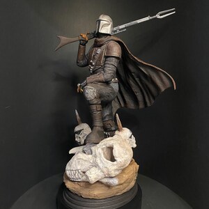 May include: A detailed figurine of The Mandalorian, clad in armor and a cape, holding a rifle. The figure stands atop a base with skulls and a black circular stand. The color palette includes silver, brown, and black.