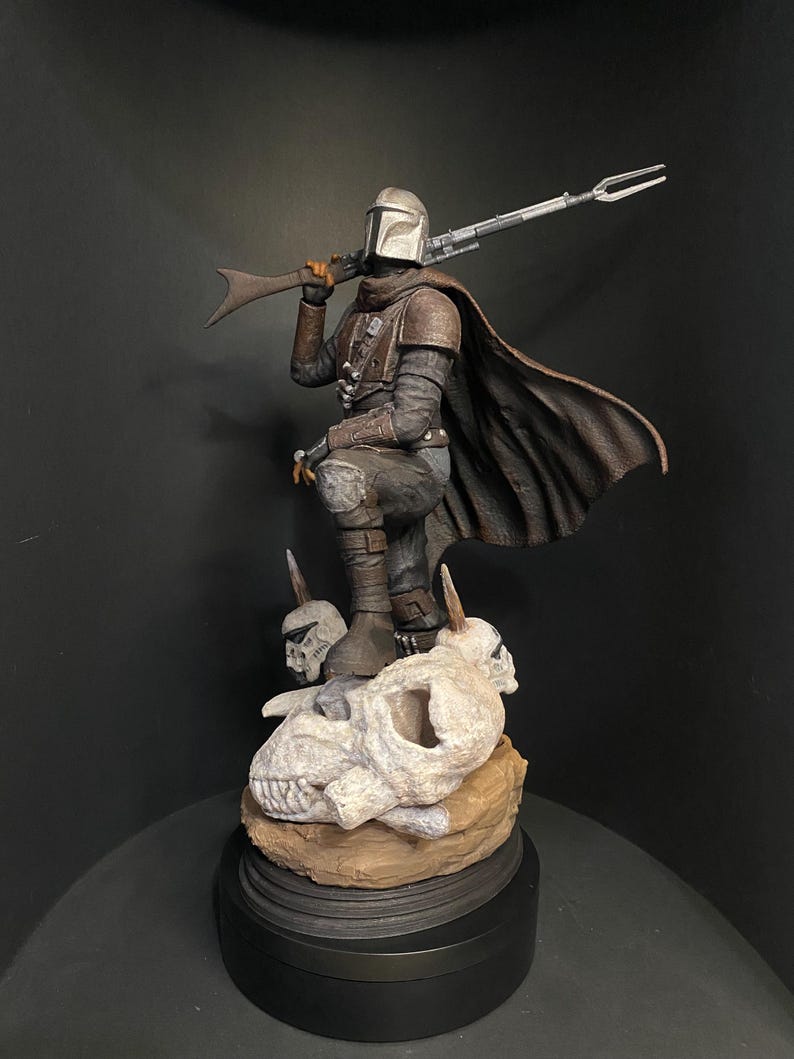 May include: A detailed figurine of The Mandalorian, clad in armor and a cape, holding a rifle. The figure stands atop a base with skulls and a black circular stand. The color palette includes silver, brown, and black.