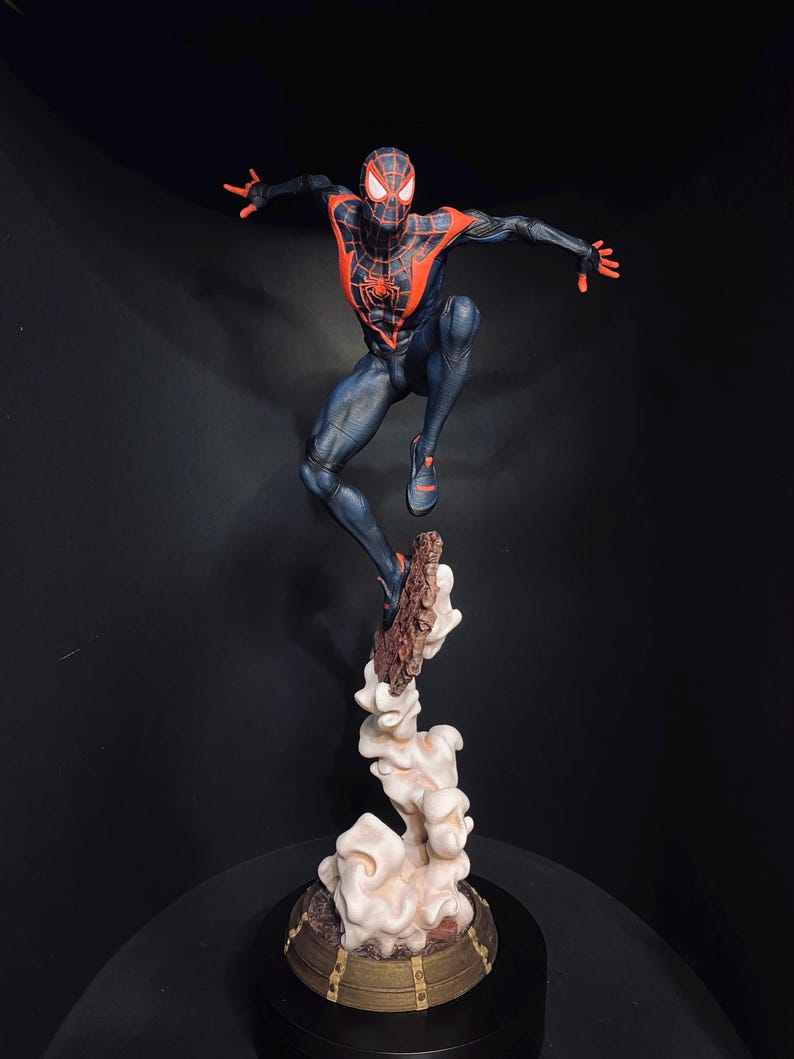 Puede incluir: Una detallada figura de Spider-Man en una pose din&aacute;mica, saltando desde una base en forma de nube. La figura es predominantemente azul marino con acentos rojos, incluyendo el emblema de la ara&ntilde;a en el pecho. La base es una mezcla de marr&oacute;n y crema.