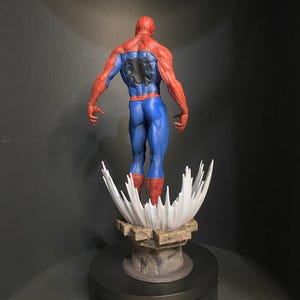 Pode incluir: Uma figura detalhada do Homem-Aranha, vista por tr&aacute;s, mostrando seu ic&ocirc;nico traje vermelho e azul. A figura est&aacute; em cima de uma base estilizada com um design branco em forma de respingo e uma plataforma rochosa marrom. A figura est&aacute; em uma base preta girat&oacute;ria.