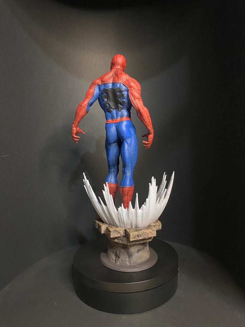 Pode incluir: Uma figura detalhada do Homem-Aranha, vista por tr&aacute;s, mostrando seu ic&ocirc;nico traje vermelho e azul. A figura est&aacute; em cima de uma base estilizada com um design branco em forma de respingo e uma plataforma rochosa marrom. A figura est&aacute; em uma base preta girat&oacute;ria.