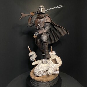 May include: A detailed figurine of The Mandalorian, clad in weathered armor, holding a rifle. The figure stands atop a sculpted base featuring a skull and two Stormtrooper helmets. The color palette includes shades of brown, gray, and black.
