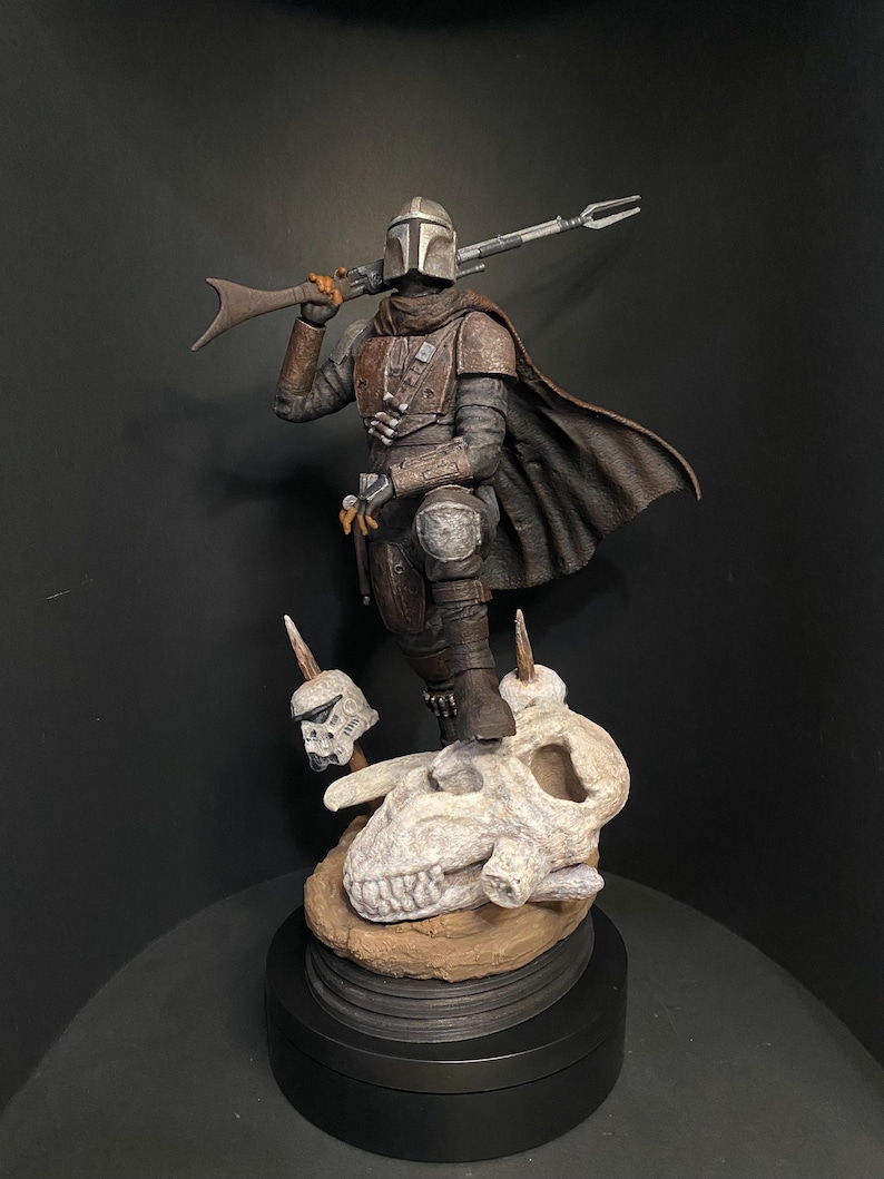 May include: A detailed figurine of The Mandalorian, clad in weathered armor, holding a rifle. The figure stands atop a sculpted base featuring a skull and two Stormtrooper helmets. The color palette includes shades of brown, gray, and black.