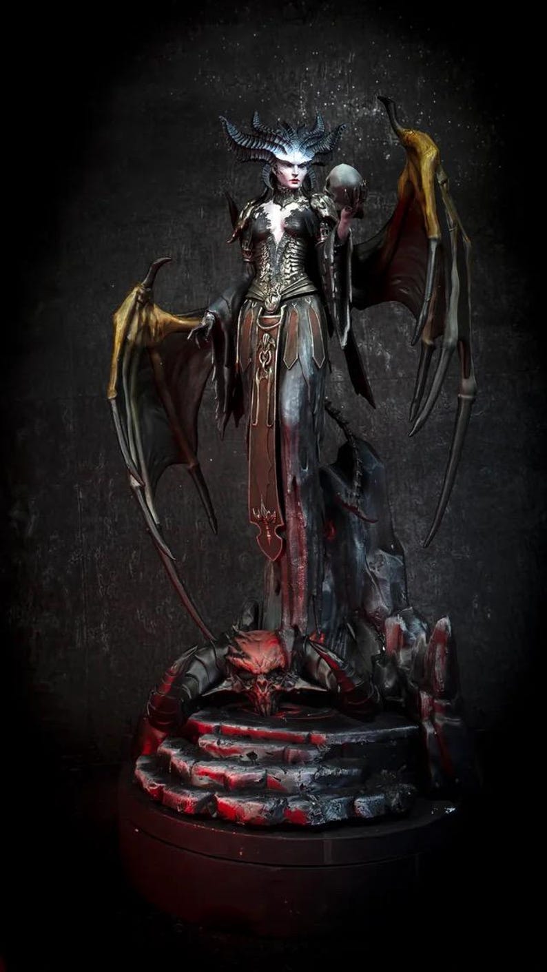 May include: A detailed figurine of a female demon with large wings, horns, and a skull. The figure is dressed in a dark gown with gold accents, standing on a stone base with a red skull.