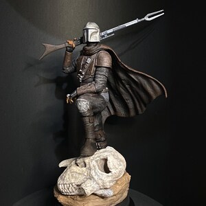 May include: A detailed figurine of a character from The Mandalorian. The figure is clad in armor, a helmet, and a dark brown cape, holding a rifle. It stands on a base with a skull and a brown rock-like structure. The background is dark.