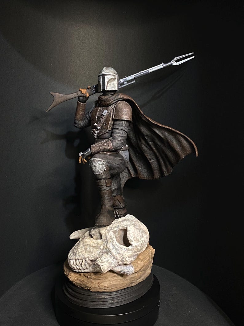 May include: A detailed figurine of a character from The Mandalorian. The figure is clad in armor, a helmet, and a dark brown cape, holding a rifle. It stands on a base with a skull and a brown rock-like structure. The background is dark.