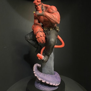 May include: A red Hellboy statue with a muscular physique, horns, and a tail. He is seated on a stone pillar, with a purple tentacle base. The figure is wearing black trousers and a belt. The statue is a detailed representation of the comic book character.