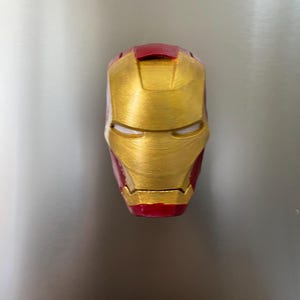 May include: A gold and red Iron Man mask, a superhero helmet, is displayed against a neutral background. The mask features a metallic gold finish with red accents on the top and bottom, and eye slits.