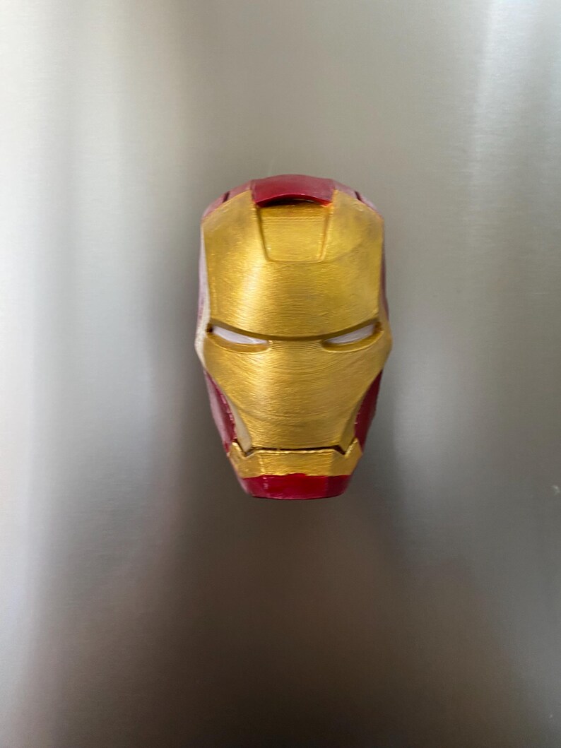 May include: A gold and red Iron Man mask, a superhero helmet, is displayed against a neutral background. The mask features a metallic gold finish with red accents on the top and bottom, and eye slits.