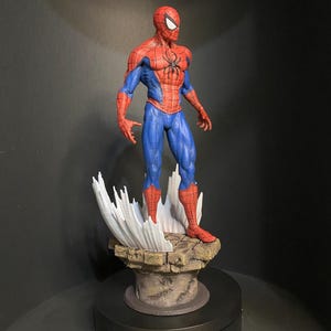 Pode incluir: Uma figura detalhada do Homem-Aranha numa pose cl&aacute;ssica. A figura &eacute; predominantemente vermelha e azul, com um emblema de aranha preta no peito. Est&aacute; sobre uma base rochosa com elementos brancos estilizados em forma de teia.