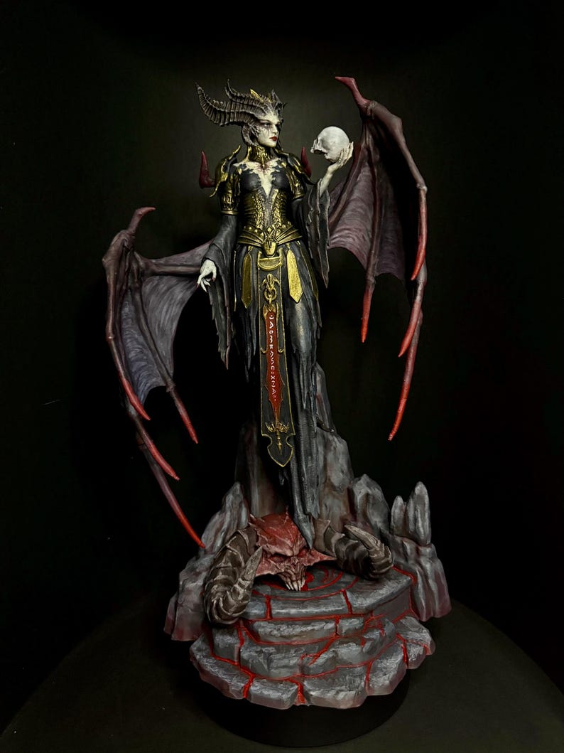 May include: A detailed figurine of a demonic figure holding a skull. The figure wears a black and gold gown, has large wings, and stands on a rocky base. The wings and base have red accents, and the background is dark.