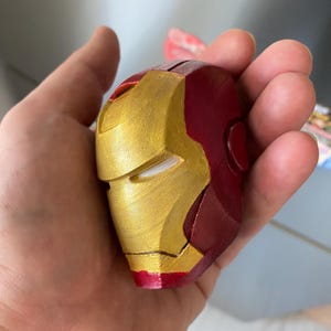 May include: A miniature Iron Man helmet, painted in metallic gold and deep red. The helmet's design is a recognizable replica of the superhero's mask, with distinct facial features and a smooth finish. The item is held in a hand.