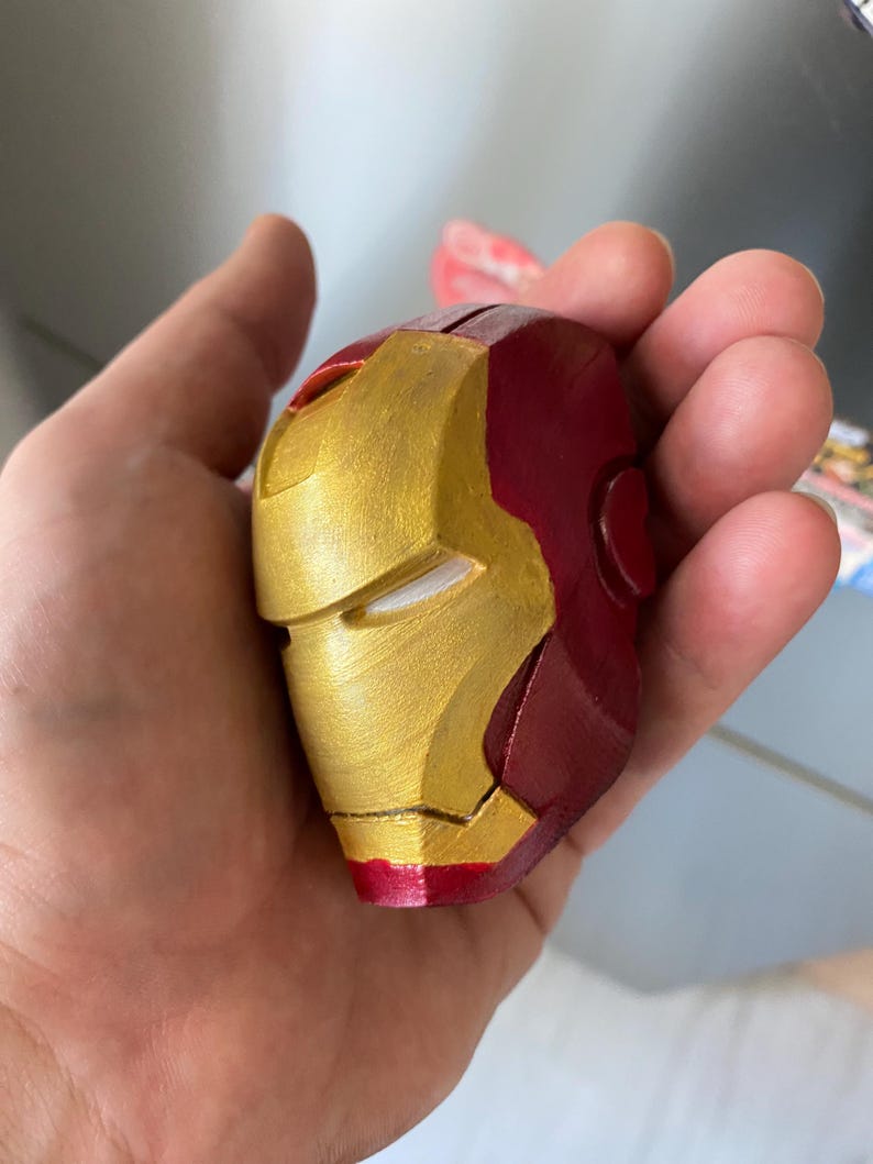 May include: A miniature Iron Man helmet, painted in metallic gold and deep red. The helmet's design is a recognizable replica of the superhero's mask, with distinct facial features and a smooth finish. The item is held in a hand.