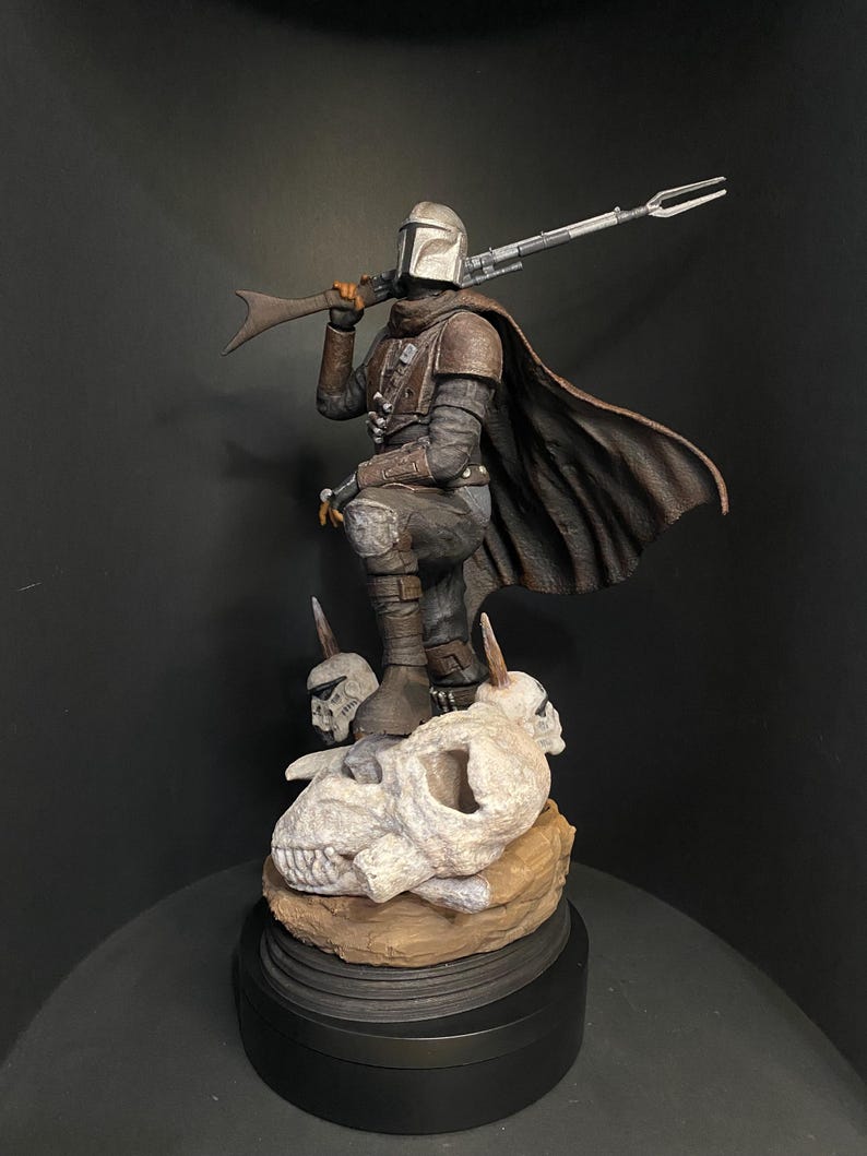 May include: A detailed figurine of The Mandalorian, a character from the Star Wars franchise, in a kneeling pose. The figure is clad in armor and a cape, holding a rifle. It stands atop a base with skulls and a brown, textured surface.