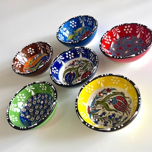 Op de afbeelding: Een verzameling van zes kleine, ovaalvormige keramische kommen. Elke kom heeft een andere levendige kleur, waaronder blauw, rood, groen, geel en bruin. De kommen zijn versierd met ingewikkelde bloemen- en geometrische patronen, met een zwarte rand en witte accenten.