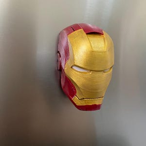 May include: A red and gold Iron Man mask, a superhero helmet, is displayed. The mask is painted with a metallic gold finish on the face and a deep red on the top and sides. The mask is a recognizable symbol from the Marvel comics.