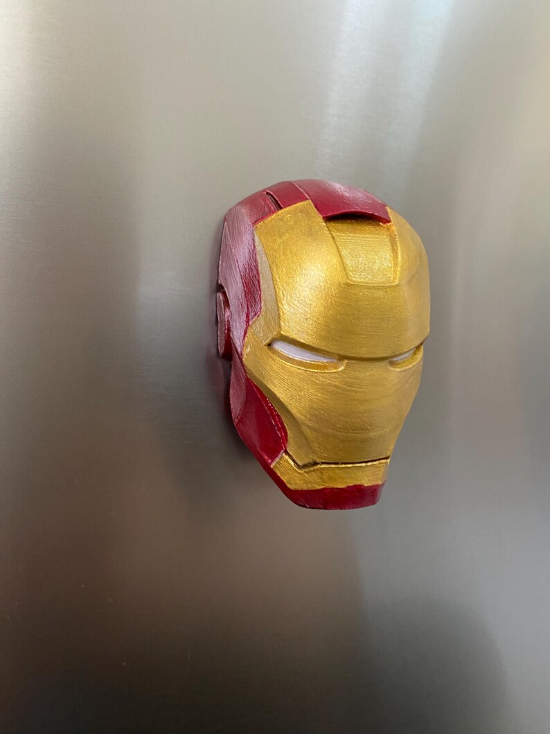 May include: A red and gold Iron Man mask, a superhero helmet, is displayed. The mask is painted with a metallic gold finish on the face and a deep red on the top and sides. The mask is a recognizable symbol from the Marvel comics.