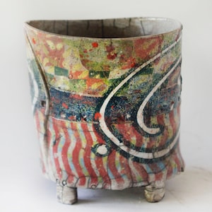 May include: A cylindrical ceramic vessel with three small feet, featuring a crackled glaze finish. The surface is decorated with a colorful pattern of red and blue wavy stripes, dark blue swirls, and speckled green, yellow, and red accents.