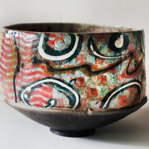 May include: A ceramic bowl featuring a dark charcoal base and a vibrant upper section decorated with red, white, and black abstract patterns, including wavy lines, circular motifs, and a multi-colored mosaic-like texture across the surface.