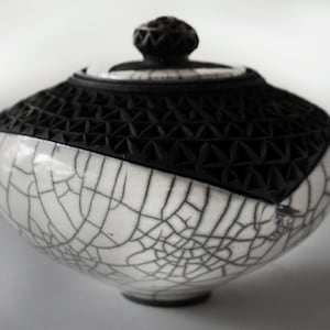 May include: A round ceramic lidded jar featuring a white base with a black crackle glaze pattern. The upper section and the lid are finished in matte black with a carved geometric triangular texture. A small knob sits atop the lid.