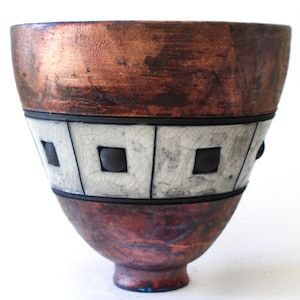 May include: A handmade ceramic bowl with a copper-coloured rim and base. The middle section has a white, cracked glaze design with black square accents. The bowl has a unique, textured appearance and a small foot.