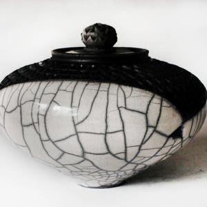 May include: A ceramic jar with a black lid and a white body. The white portion of the jar is covered in a network of dark gray lines, creating a cracked effect. The lid has a textured, woven appearance.