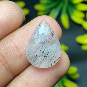 May include: A clear, teardrop-shaped gemstone with black needle-like inclusions. The polished stone is held between fingers, showcasing its translucent quality and the intricate patterns within. The gem is a natural mineral.