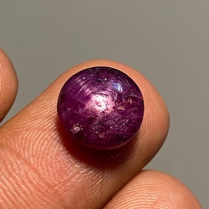 Pode incluir: Uma gema de rubi polida e redonda, exibindo um tom vermelho-púrpura profundo. A gema exibe um efeito de estrela, com a luz refletindo em sua superfície. A pedra é segurada entre os dedos, mostrando seu tamanho e cor.