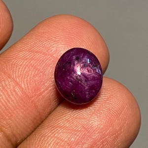 May include: A polished, oval-shaped gemstone with a deep magenta hue. The surface displays a mix of dark and light inclusions, creating a textured appearance. The gem is held between fingers, showcasing its size and color.